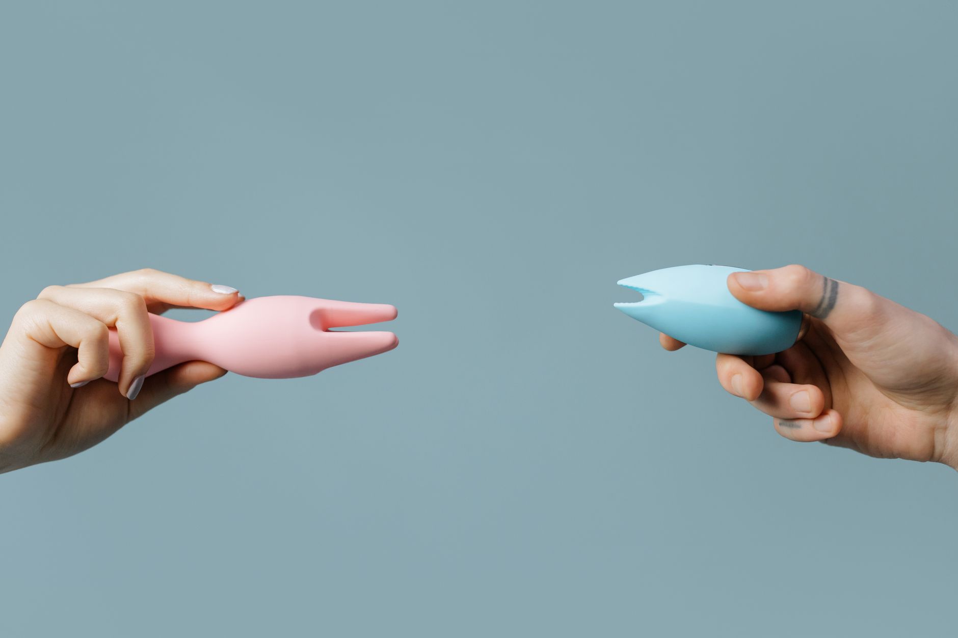 Two hands holding yellow and blue silicone vibrators against a pastel background.