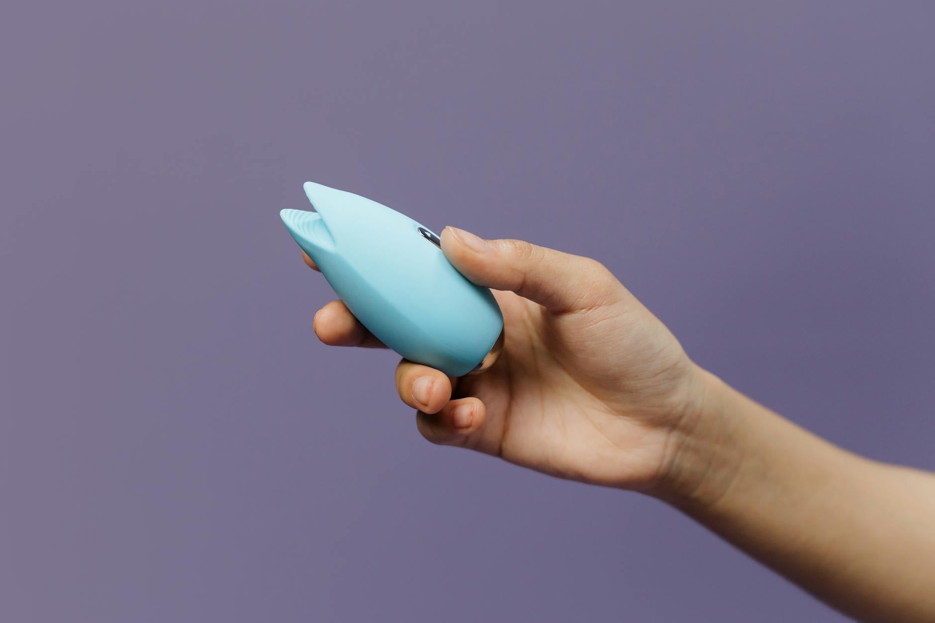 Eine Hand hält einen blauen Silikonvibrator vor violettem Hintergrund, symbolisierend Selbstliebe und weibliche Sexualität