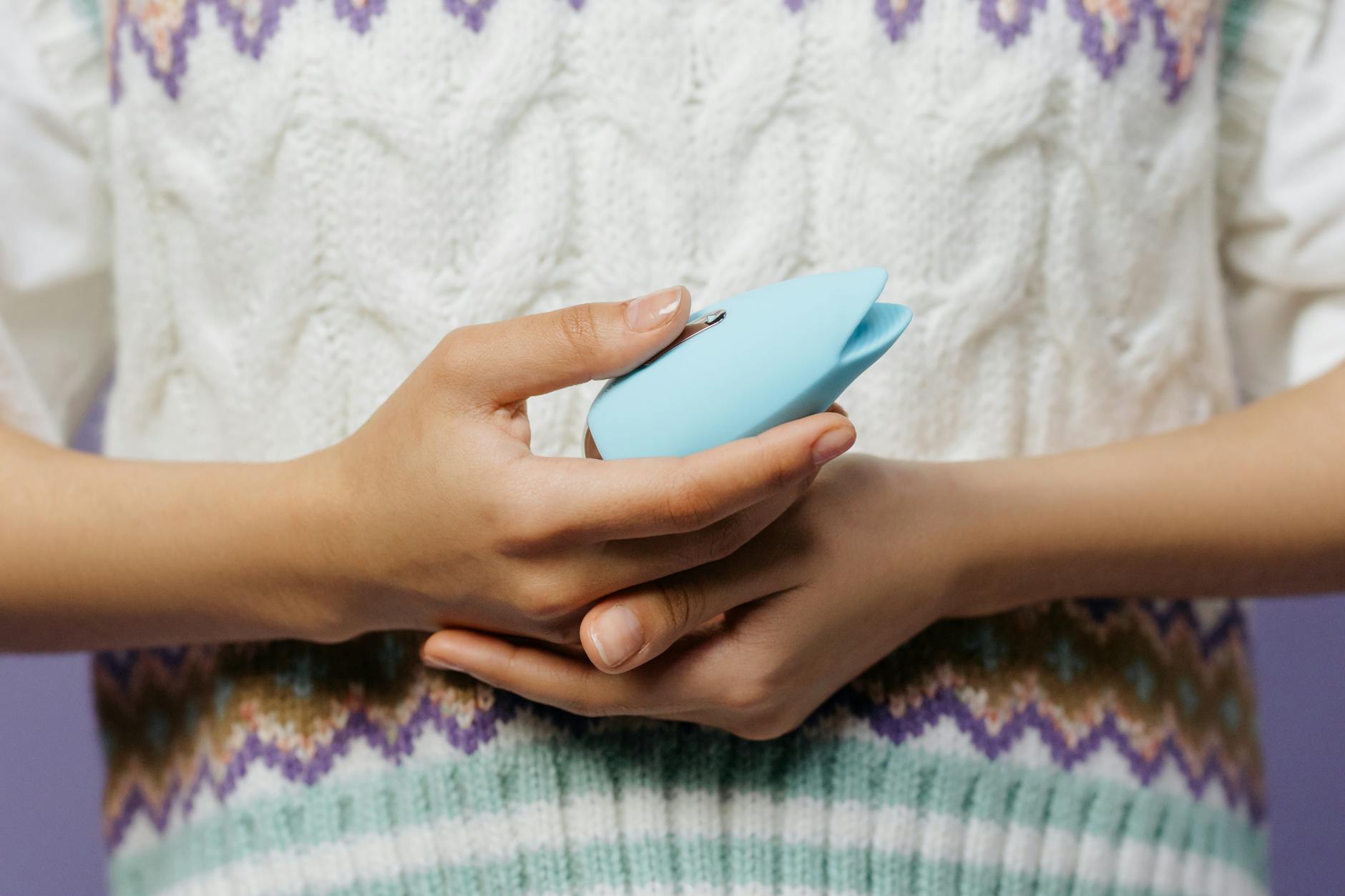 Close-up of hands holding a blue clitoral vibrator against a knitted sweater.